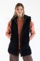 A woman modeling a black hooded zip-front vest over a terracotta long-sleeve top and striped pants, photographed against a plain white studio background.