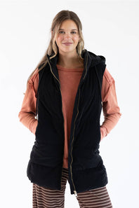 A woman modeling a black hooded zip-front vest over a terracotta long-sleeve top and striped pants, photographed against a plain white studio background.