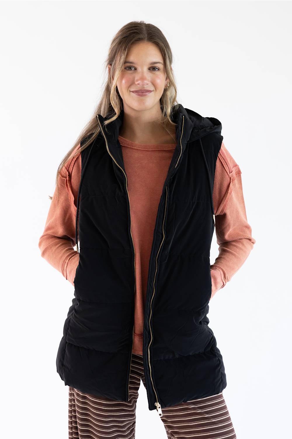 A woman modeling a black hooded zip-front vest over a terracotta long-sleeve top and striped pants, photographed against a plain white studio background.