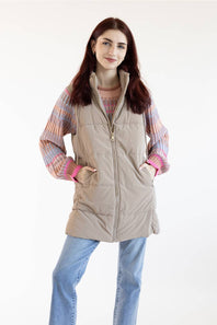 A woman modeling a light beige sleeveless puffer vest over a pink and orange striped knit sweater, paired with light blue jeans, against a plain white background.