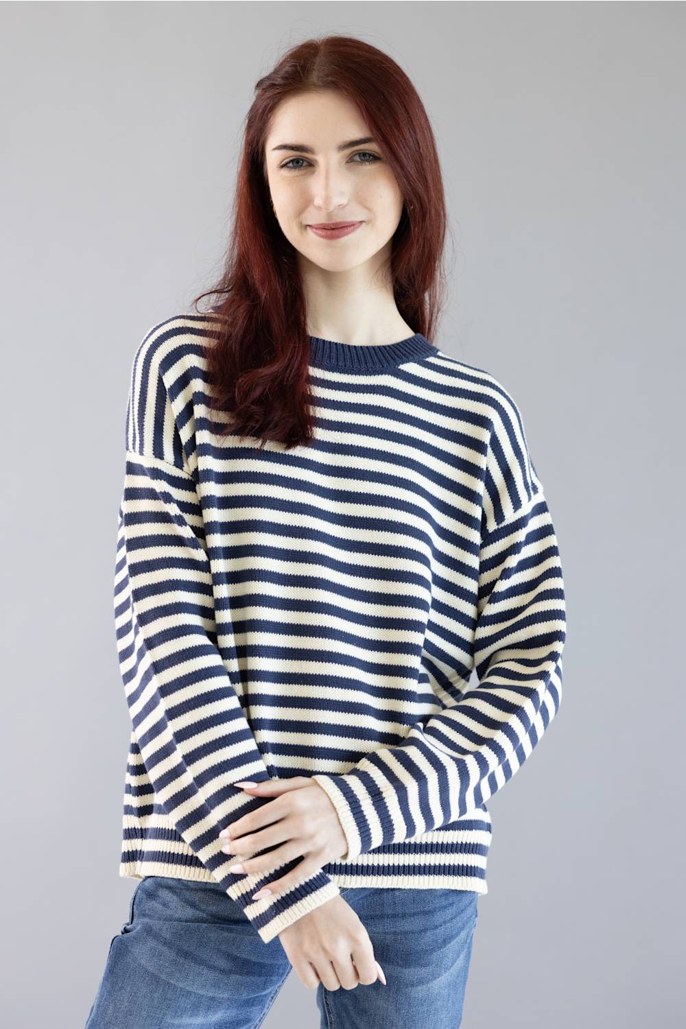 A woman with red hair wearing a navy and cream striped knit sweater and blue jeans, standing against a solid gray studio background.