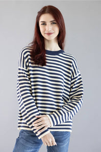 A woman with red hair wearing a navy and cream striped knit sweater and blue jeans, standing against a solid gray studio background.
