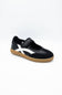A black slip-on shoe with a white side stripe and a brown gum sole, photographed on a clean white background.