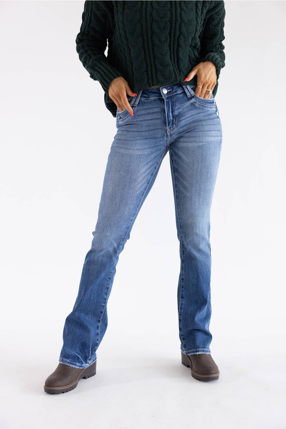 A person wearing medium-wash blue jeans, a dark green cable-knit sweater, and brown ankle boots, standing against a plain white studio background.