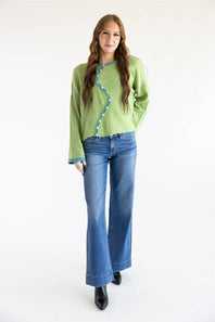 A woman modeling a light green knit sweater with a wavy teal trim detail down the front, paired with blue jeans and black boots, against a plain white studio background.