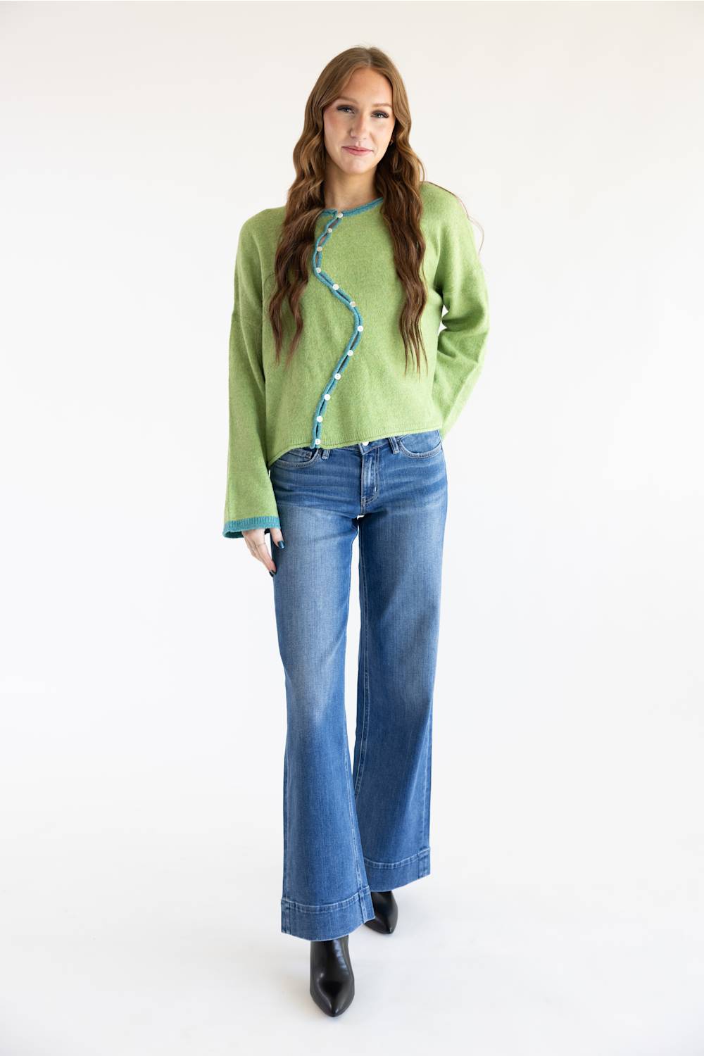 A woman modeling a light green knit sweater with a wavy teal trim detail down the front, paired with blue jeans and black boots, against a plain white studio background.