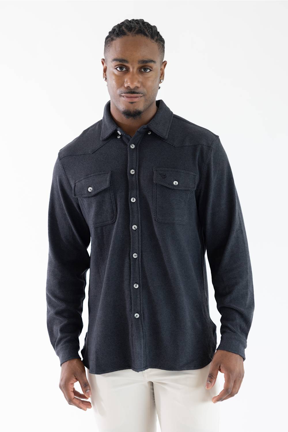 A man wearing a dark navy long-sleeve western-style button-up shirt with two chest pockets, paired with off-white pants, photographed against a plain white studio background.