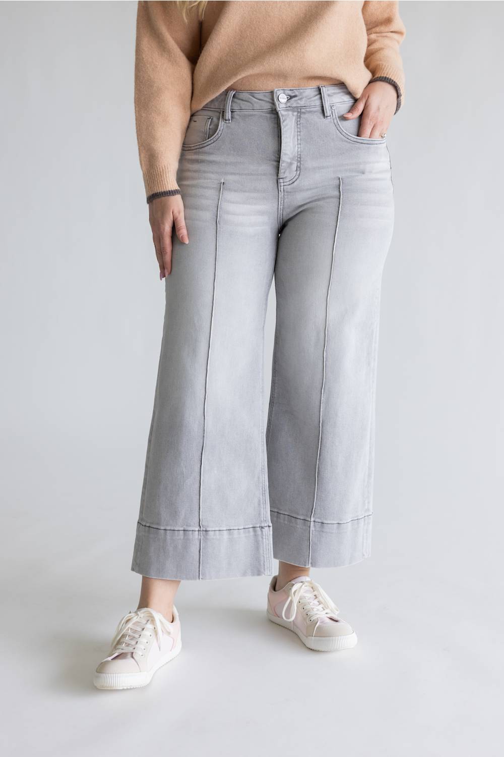 A person wearing light gray wide-leg cropped jeans with rolled hems, paired with a beige sweater and white sneakers, standing against a light gray studio background.