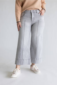 A person wearing light gray wide-leg cropped jeans with rolled hems, paired with a beige sweater and white sneakers, standing against a light gray studio background.