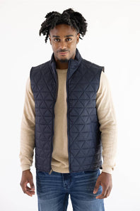 A man wearing a dark navy blue diamond-quilted puffer vest over a beige long-sleeve shirt and blue jeans, standing against a plain white background.