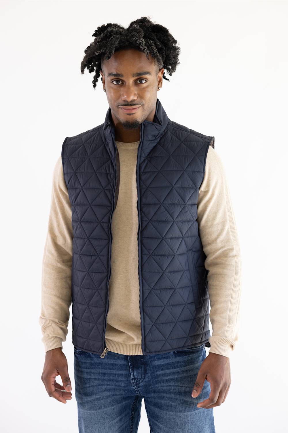 A man wearing a dark navy blue diamond-quilted puffer vest over a beige long-sleeve shirt and blue jeans, standing against a plain white background.