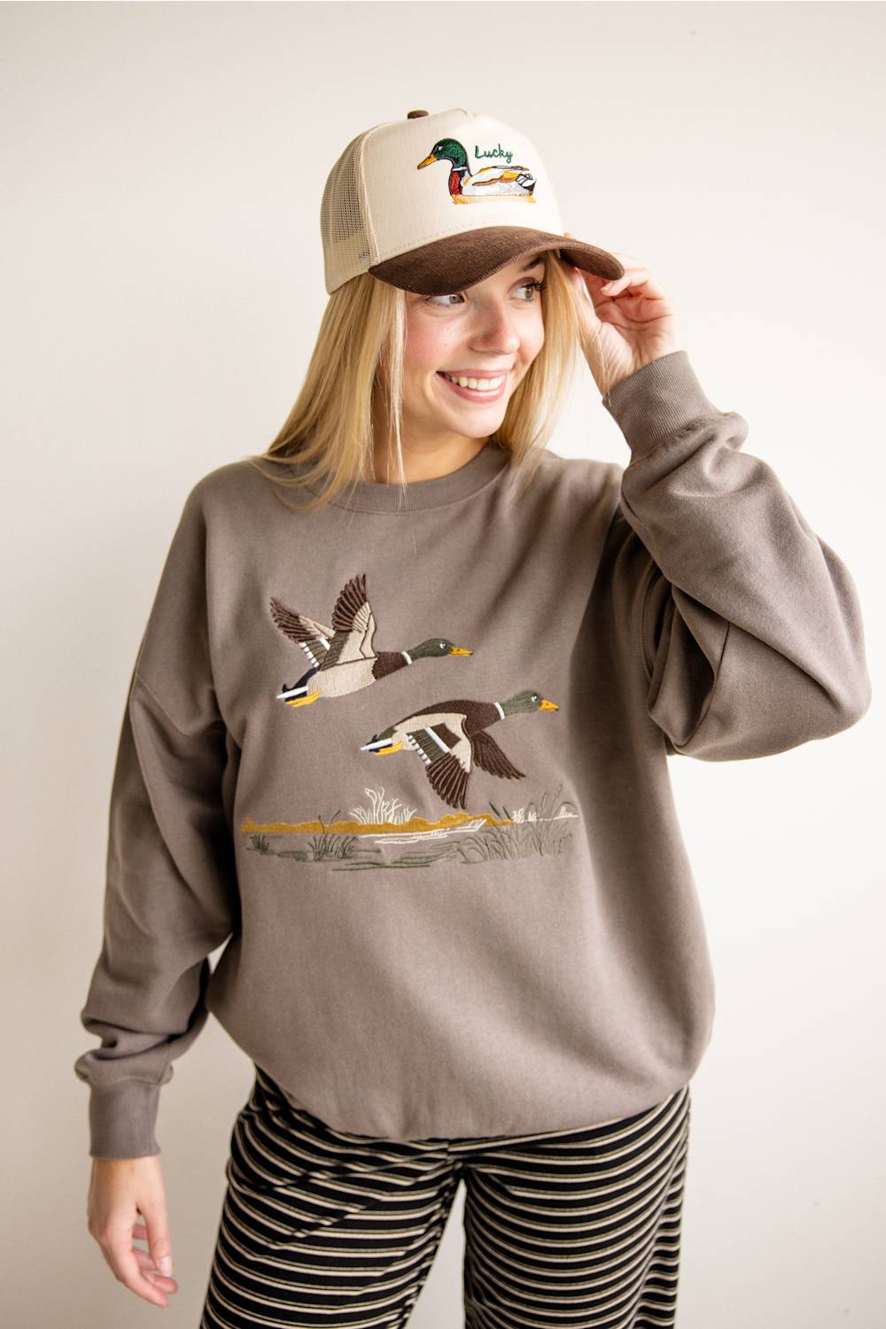 A woman wearing a taupe crewneck sweatshirt with a detailed embroidered mallard duck graphic, paired with black and white striped pants and a beige trucker hat with a duck logo.