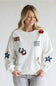 A woman wearing a white crewneck sweatshirt decorated with various embroidered patches, including a cowboy patch, a horseshoe, a rose, and blue stars, paired with light gray jeans against a plain light gray background.
