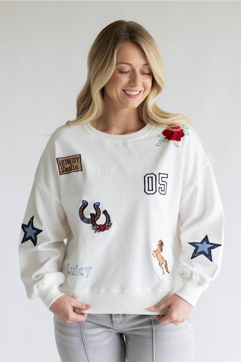 A woman wearing a white crewneck sweatshirt decorated with various embroidered patches, including a cowboy patch, a horseshoe, a rose, and blue stars, paired with light gray jeans against a plain light gray background.