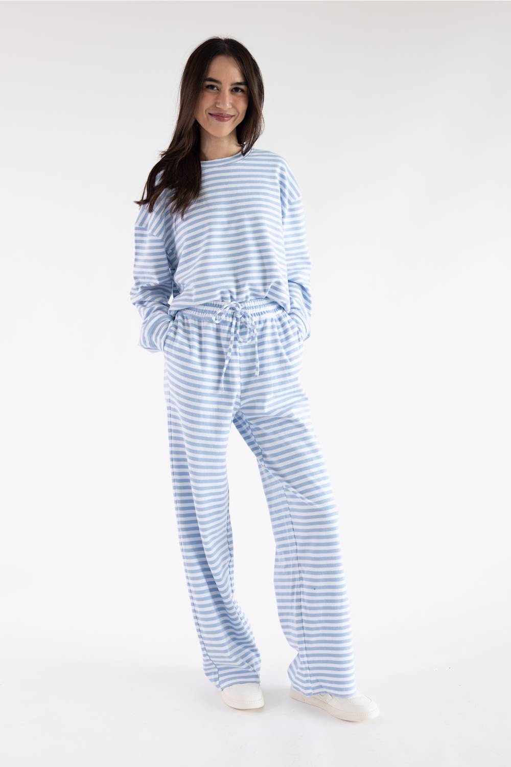 A woman modeling a light blue and white striped long-sleeve jumpsuit with a drawstring waist, photographed against a plain white studio background.