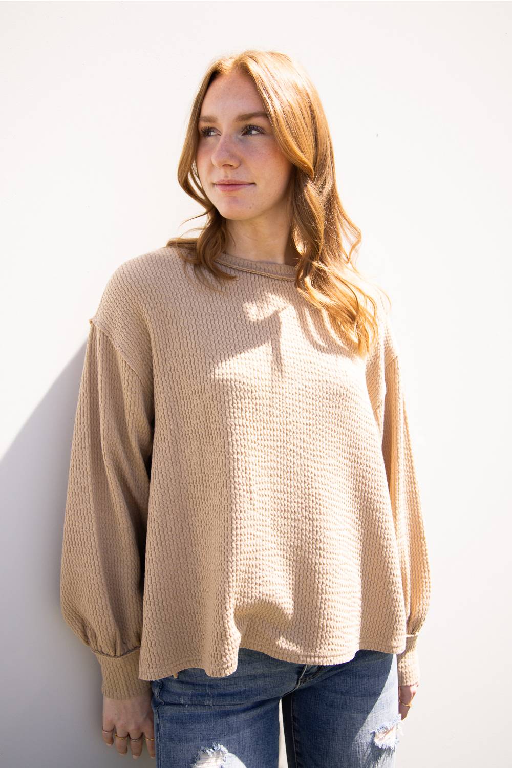 A woman with long light brown hair wearing a beige waffle-knit long-sleeve sweater and blue jeans, standing against a white wall.