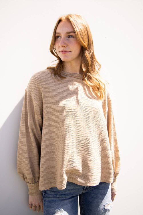 A woman with long light brown hair wearing a beige waffle-knit long-sleeve sweater and blue jeans, standing against a white wall.