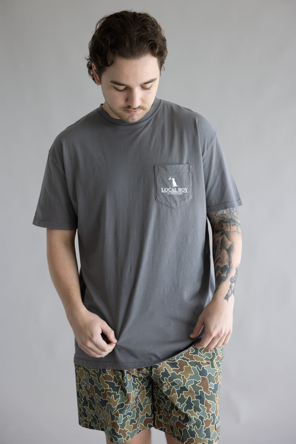 Local Boy Vintage Emblem T-Shirt for Men in Grey; Man wearing a gray t-shirt with a pocket and patterned shorts on a plain background