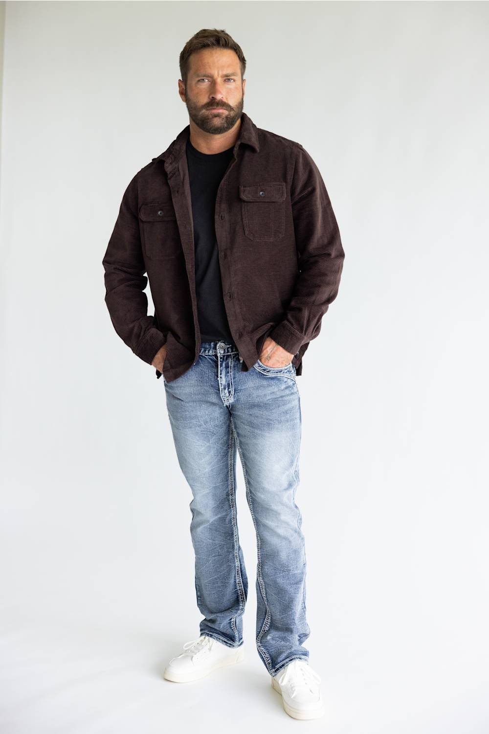 A man wearing a dark brown corduroy jacket over a black t-shirt, paired with light blue jeans and white sneakers, standing against a plain white studio background.