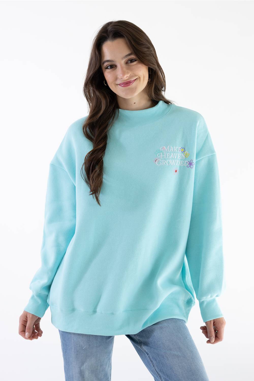 A woman wearing a light aqua blue crewneck sweatshirt with a colorful graphic on the left chest that reads  paired with light wash blue jeans, against a plain white background.