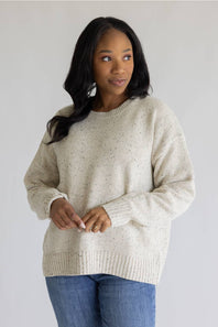A woman wearing a cream-colored, speckled knit crewneck sweater and blue jeans, standing against a plain light gray background.