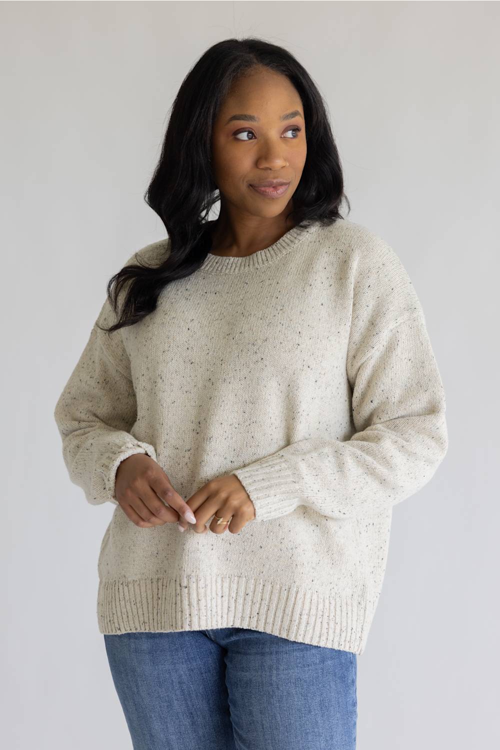 A woman wearing a cream-colored, speckled knit crewneck sweater and blue jeans, standing against a plain light gray background.