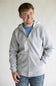 A smiling man wearing a light gray zip-up hoodie with a small yellow Carhartt logo patch on the pocket, layered over a blue t-shirt and paired with blue jeans, against a plain light gray background.