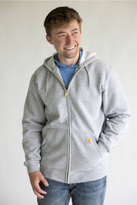 A smiling man wearing a light gray zip-up hoodie with a small yellow Carhartt logo patch on the pocket, layered over a blue t-shirt and paired with blue jeans, against a plain light gray background.