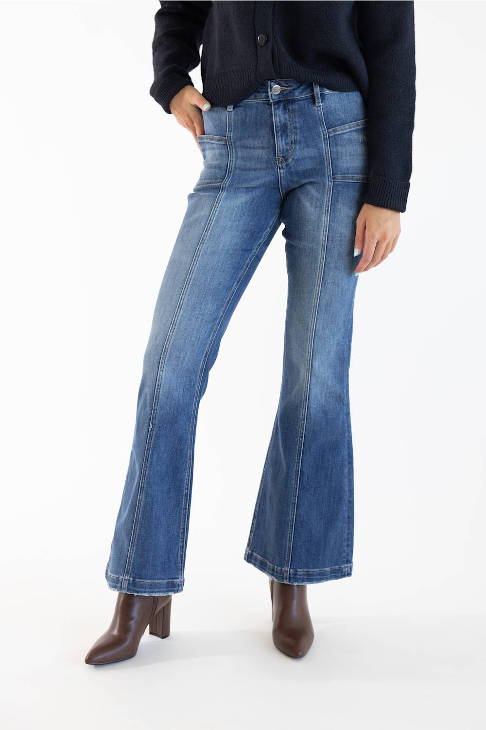 A model wearing a pair of medium-dark -wash blue denim flare jeans, styled with a dark navy cardigan and brown leather ankle boots, shot against a plain white studio background.