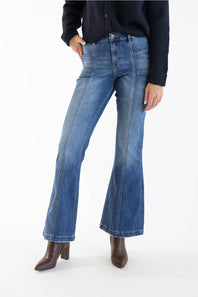 A model wearing a pair of medium-dark -wash blue denim flare jeans, styled with a dark navy cardigan and brown leather ankle boots, shot against a plain white studio background.