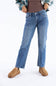 A person wearing medium-wash blue straight-leg jeans, a brown ribbed top, a green and white plaid flannel shirt, and tan suede slip-on shoes, shown from the waist down on a white studio background.