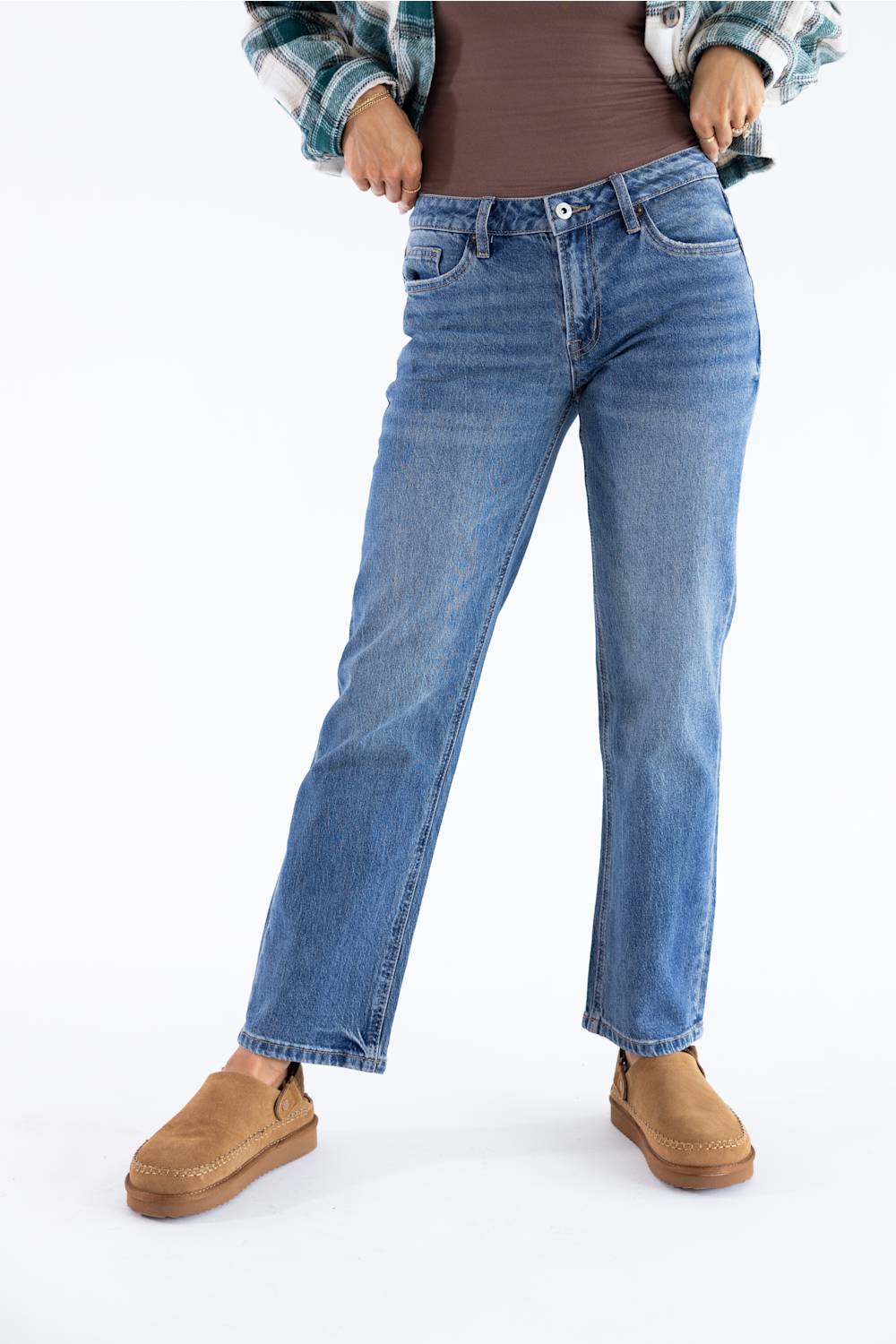 A person wearing medium-wash blue straight-leg jeans, a brown ribbed top, a green and white plaid flannel shirt, and tan suede slip-on shoes, shown from the waist down on a white studio background.