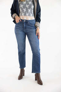 A model wearing a pair of dark-wash blue straight-leg jeans, styled with a white lace top, a dark navy jacket, and brown ankle boots, set against a plain white studio background.