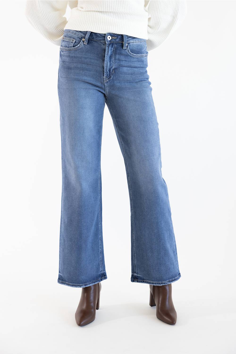 A person wearing medium-wash blue wide leg jeans with a white ribbed sweater and brown heeled ankle boots, shown from the waist down against a plain white studio background.