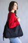 A woman wearing a red knit sweater and blue jeans carries a large black suede-like shoulder bag against a plain gray studio background.