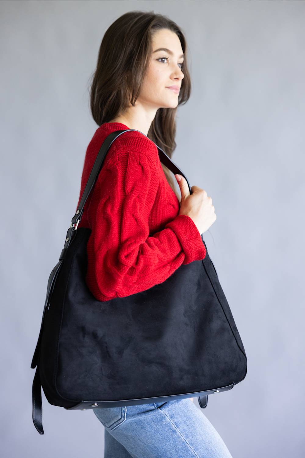 A woman wearing a red knit sweater and blue jeans carries a large black suede-like shoulder bag against a plain gray studio background.