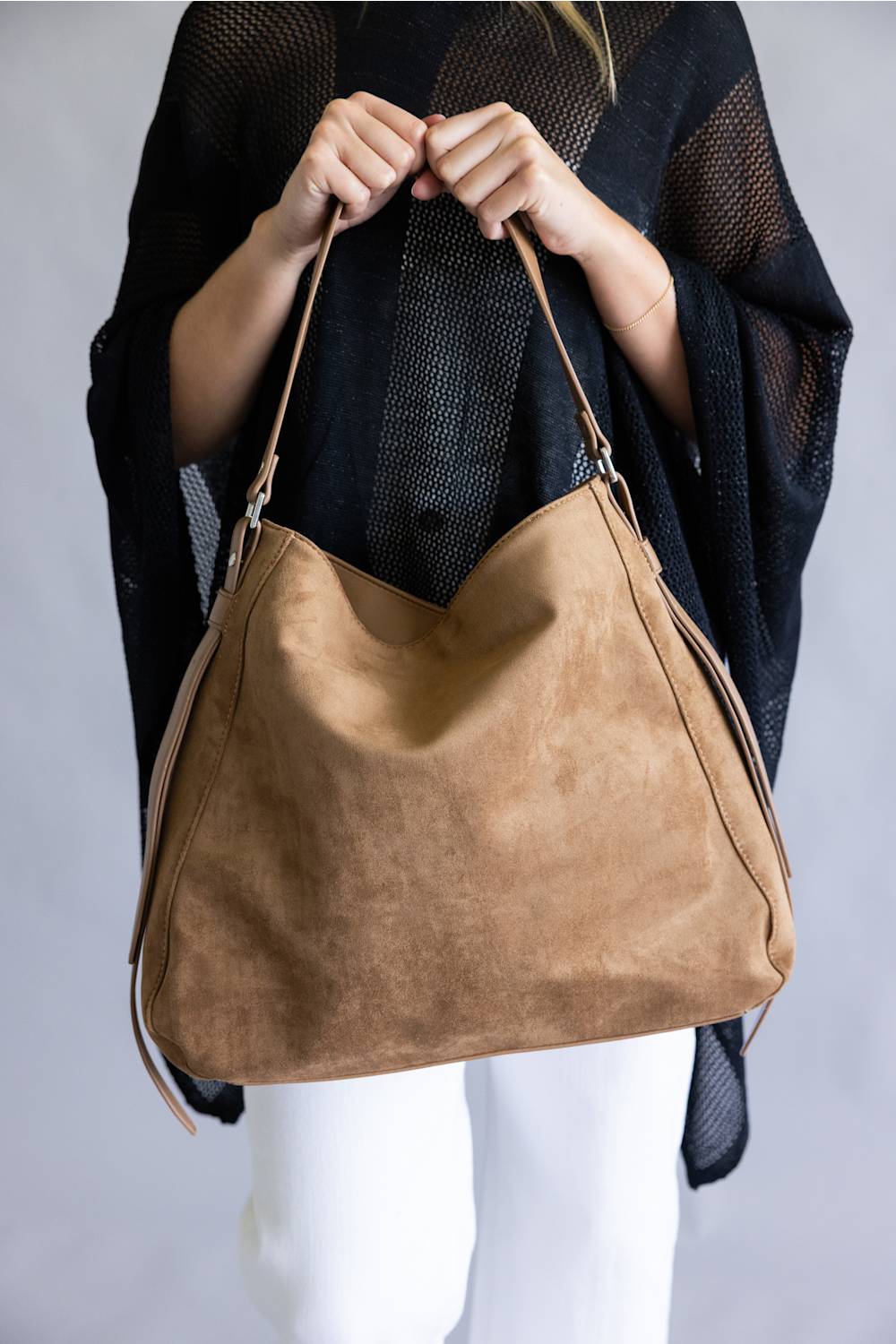 A person wearing a black open-knit cardigan and white pants holds up a large tan suede hobo shoulder bag against a light gray studio background.