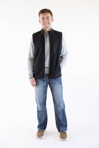 A man wearing a dark charcoal gray zippered fleece vest over a light gray zip-up sweater, blue jeans, and brown shoes, standing against a plain white studio background.