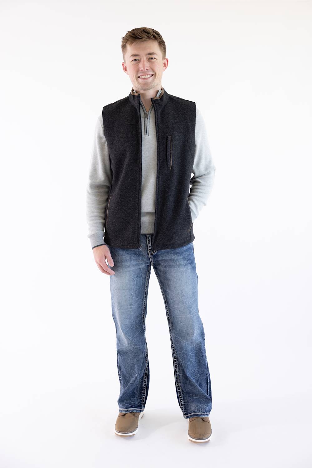 A man wearing a dark charcoal gray zippered fleece vest over a light gray zip-up sweater, blue jeans, and brown shoes, standing against a plain white studio background.