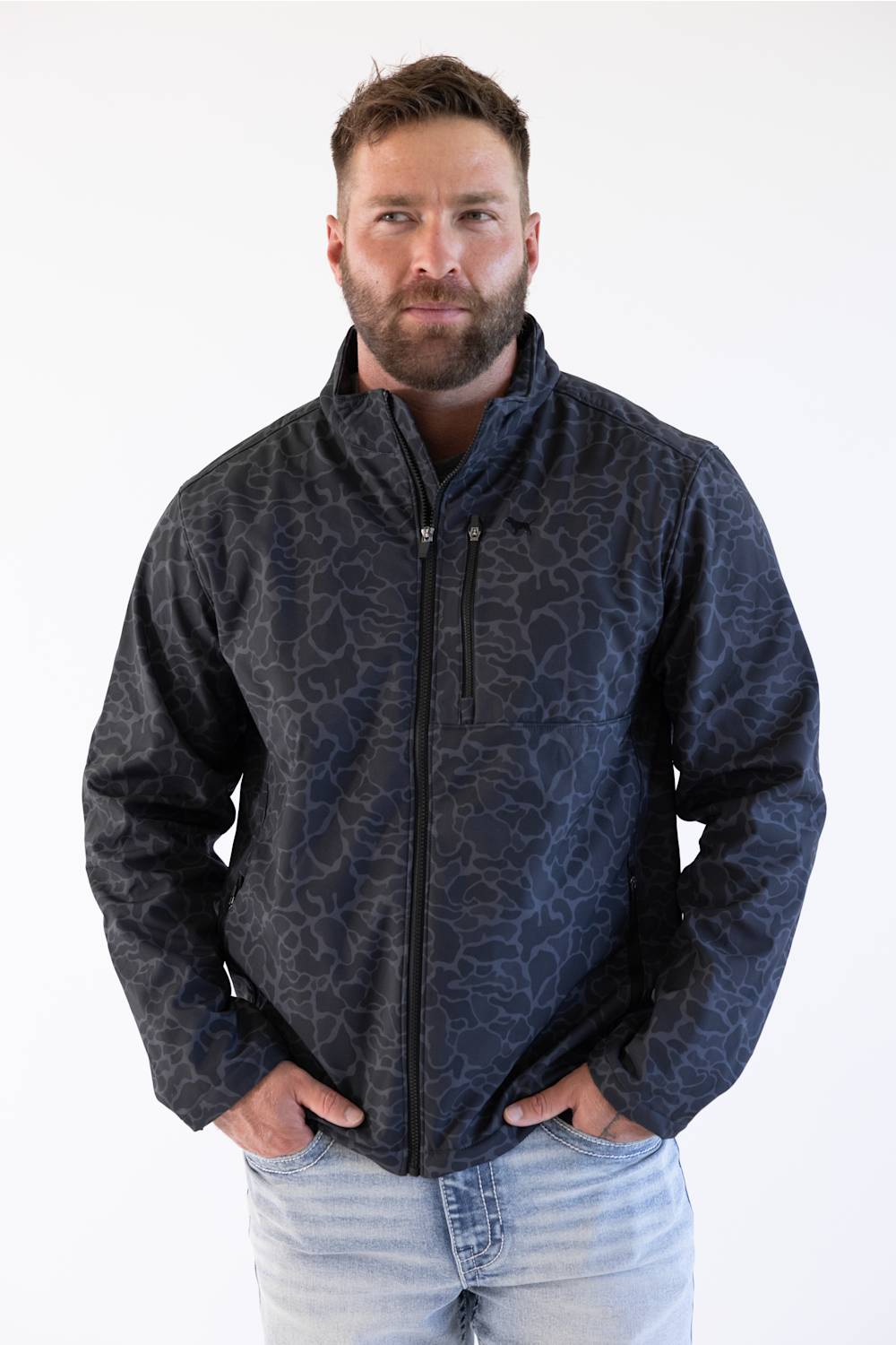 A man wearing a dark navy blue zip-up jacket with a subtle tonal abstract pattern, paired with light blue jeans, standing against a plain white background.