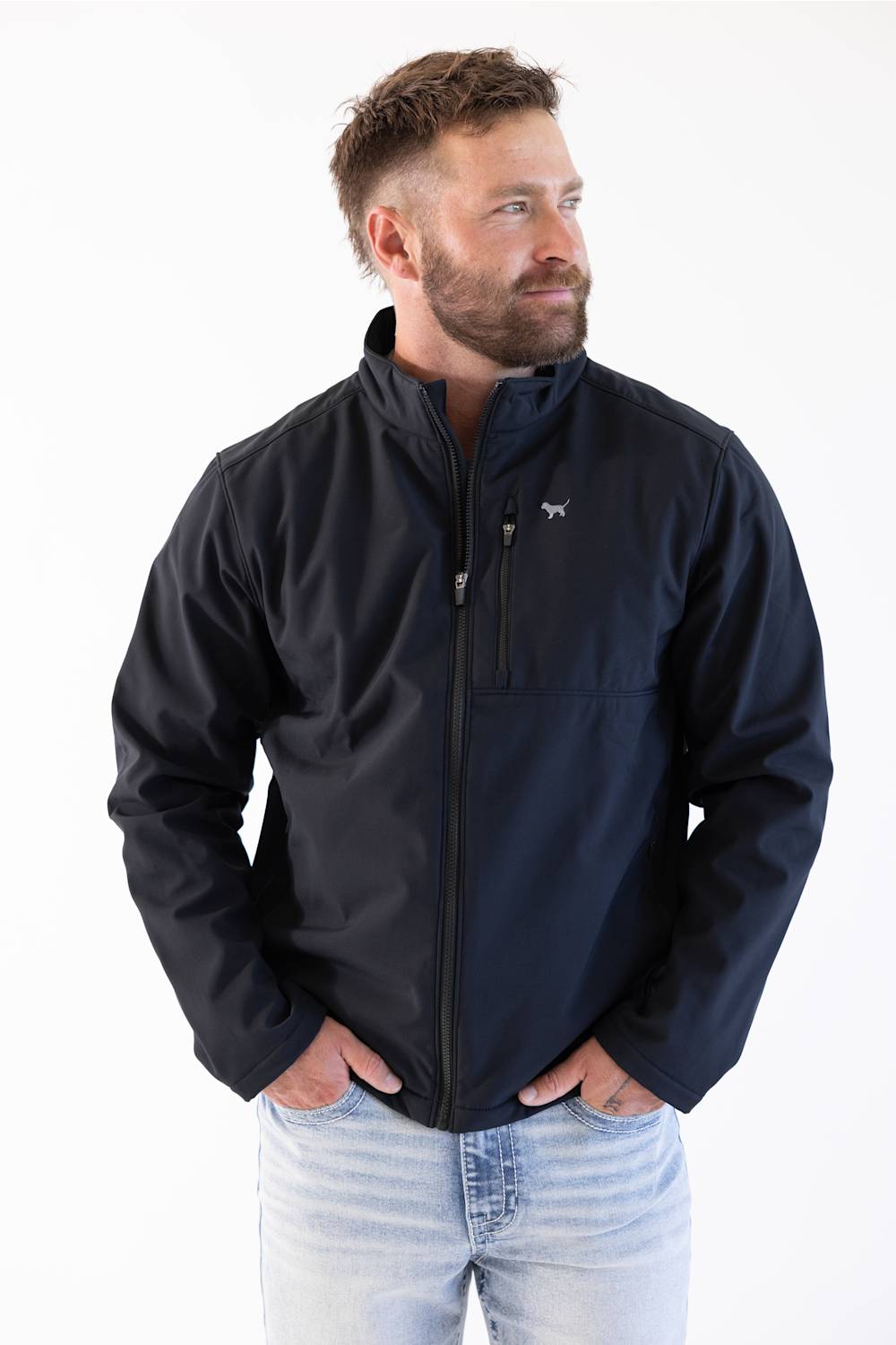A man wearing a dark navy blue zip-up jacket with a small white dog logo on the left chest, paired with light blue jeans, photographed against a plain white background.