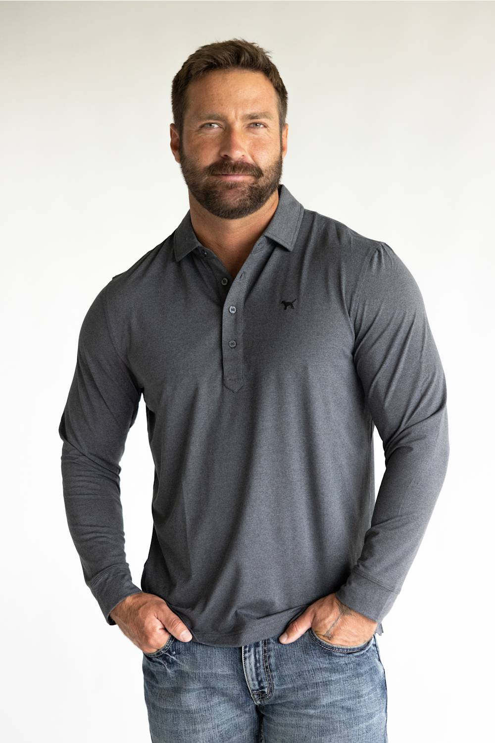 A muscular man with a beard wearing a dark gray long-sleeve polo shirt and blue jeans, standing against a plain white background.