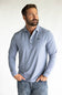A man wearing a light blue long-sleeve polo shirt with a small black embroidered logo on the chest, paired with blue jeans, standing against a plain white background.