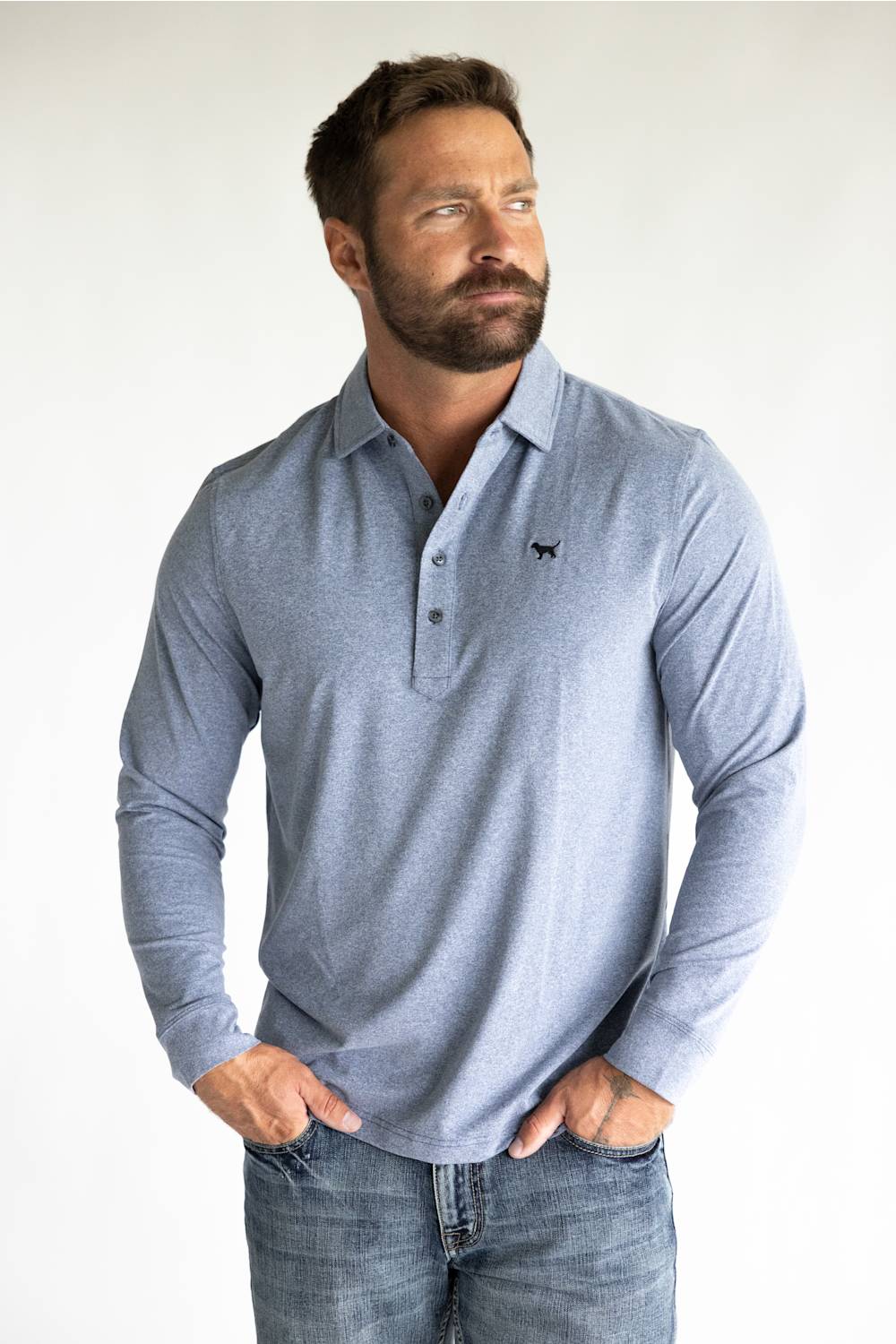 A man wearing a light blue long-sleeve polo shirt with a small black embroidered logo on the chest, paired with blue jeans, standing against a plain white background.