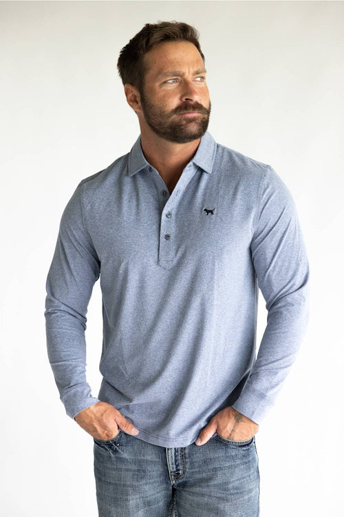 A man wearing a light blue long-sleeve polo shirt with a small black embroidered logo on the chest, paired with blue jeans, standing against a plain white background.