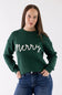 A woman wearing a dark green crewneck sweatshirt with 'Merry' written in white cursive across the chest, paired with blue jeans, against a plain white studio background.