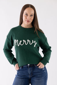 A woman wearing a dark green crewneck sweatshirt with 'Merry' written in white cursive across the chest, paired with blue jeans, against a plain white studio background.