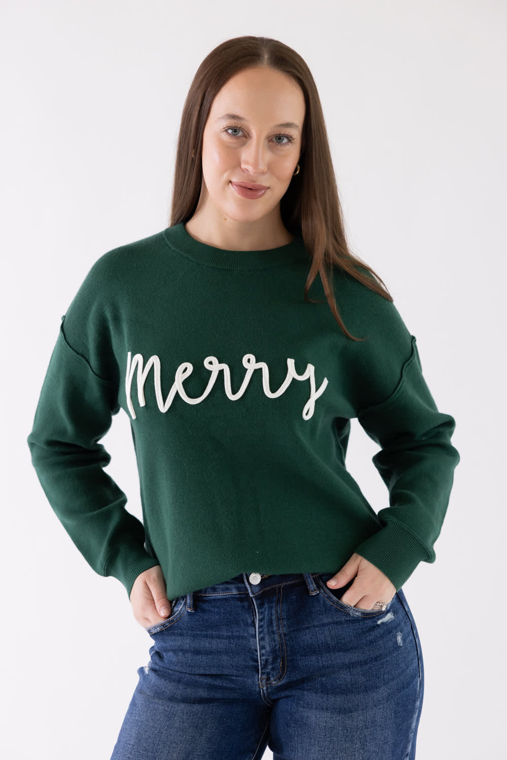 A woman wearing a dark green crewneck sweatshirt with 'Merry' written in white cursive across the chest, paired with blue jeans, against a plain white studio background.