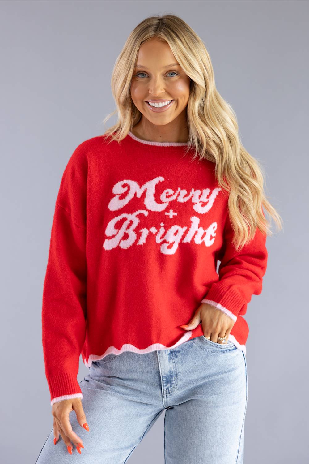 A smiling blonde woman wearing a bright red knit sweater with 'Merry Bright' in white script across the front, paired with light blue jeans, against a solid gray studio background.