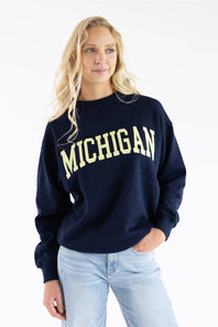 A woman wearing a navy blue crewneck sweatshirt with 'MICHIGAN' printed in cream across the chest, paired with light wash blue jeans, against a plain white background.